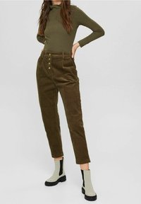 Femme portant un haut côtelé à manches longues vert olive, un pantalon en velours côtelé marron taille haute avec des boutons, et des bottines beige avec des côtés élastiques noirs.