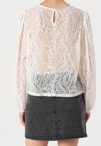 Blouse en dentelle blanche avec manches bouffantes, comportant un détail dos en forme de trou clé. Associée à une jupe en jean gris foncé ornée de clous noirs.