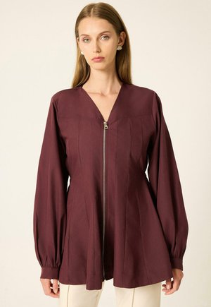 Femme portant un chemisier zippé manches longues couleur bordeaux foncé, avec un col en V et un ourlet légèrement évasé, associé à un pantalon clair.