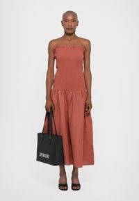 Φόρεμα χωρίς straps σε σκουριά χρώμα με υφή στον κορμό και ριγωμένο λαιμόκοψη, διαθέτοντας μια ρέουσα φούστα κα�ι κρατώντας μια μαύρη τσάντα tote που απεικονίζει "LOVE MOSCHINO."