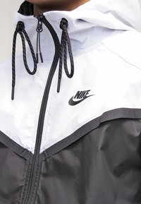 Nike vindjacka med en design i svart och vitt färgblock, med dragkedja, snoddar och en framträdande logotyp på bröstet.
