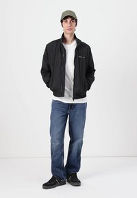 Black lightweight bomber jacket with a zip, worn over a white t-shirt. Paired with blue jeans and black sneakers, topped with a green cap.