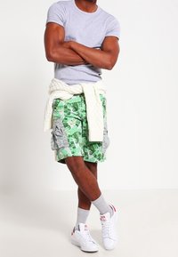 Zielone spodenki cargo w kamuflażu z wieloma kieszeniami, szary T-shirt, biały sweter przewiązany w talii oraz białe buty z czerwonymi akcentami.