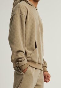 Personne portant un sweat à capuche beige texturé et un pantalon assorti, mains partiellement dans les poches, visible des épaules aux hanches, fond neutre.