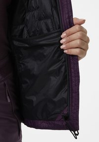 Vnitřek fialové zateplené bundy s hladkým černým podšívkou. Obsahuje kapsu na zip a nastavitelné stahování na dolním lemu.