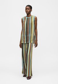 Femme portant un haut sans manches et un pantalon large à rayures verticales multicolores, debout devant un fond blanc uni.