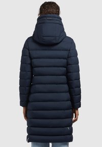 Person klädd i en lång marinblå kviltad pufferjacka med hög stoppad krage och huva, sedd bakifrån.