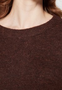 Hnědý vlnený svetr s kulatým v ýstřihem a texturovaným pleteným vzorem. Látka působí jemně a trochu chlupatě.