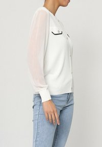 Cardigan blanc avec ourlet et poignet côtelés. Manches longues plissées et transparentes ; deux petites poches avec un bord noir. Design à boutons sur le devant.