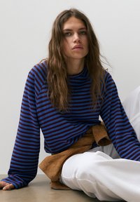 Giovane donna con lunghi capelli ondulati che indossa una maglia a maniche lunghe a righe blu e rosso scuro, pantaloni bianchi e una giacca marrone legata in vita.