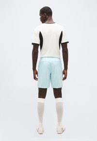 Άνδρας που στέκεται με την πλάτη προς το μέρος μας, φοράει λευκό και μαύρο μπλουζάκι με κοντά μανίκια, ανοιχτόχρωμα μπλε σορτς, λευκές κάλτσες μέχρι το γόνατο και λευκά παπούτσια ποδοσφαίρου.