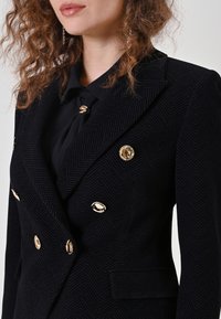 Blazer noir croisé à motif chevrons avec boutons dorés et cols à pointe. Tissu texturé et deux poches latérales.