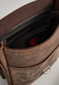 Interno di una borsa in pelle marrone, con una fodera in tessuto scuro, tasca con zip e un'etichetta del marchio. Cuciture visibili e dettagli in metallo.