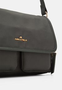 Borsa a tracolla verde scuro in materiale liscio, con logo dorato "ANNA FIELD", chiusura a patta e due tasche frontali con dettaglio cerniera.