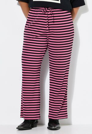Personne portant un pantalon ample à jambes larges avec des rayures horizontales noires et roses et des chaussures noires, debout devant un fond blanc.