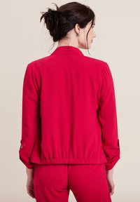 Giacca rossa leggera con orlo arricciato e maniche lunghe. Presenta un colletto e dettagli con maniche arrotolate. Tessuto liscio, senza disegni.