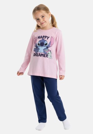 Ein junges Mädchen steht lächelnd da, trägt ein pinkfarbenes Langarmshirt mit „Happy Dreamer“ und einer Cartoon-Figur, dunkelblaue Hose und weiße Socken.
