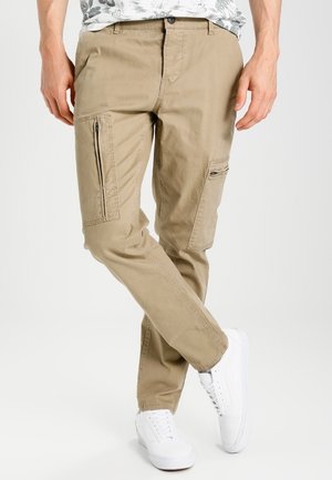 Mann trägt beige Cargohosen mit Reißverschlusstaschen und weiße Sneaker, steht mit überkreuzten Beinen vor einem schlichten Hintergrund.
