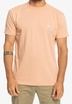 Homme portant un t-shirt pêche à manches courtes avec un petit logo blanc sur la poitrine et un pantalon kaki, debout avec les bras détendus.