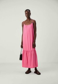 Γυναίκα ντυμένη με ροζ αμάνικο maxi φόρεμα, με μαύρες σανδάλια και κρατώντας μια μαύρη τσάντα μπροστά από απλό ανοιχτό φόντο.