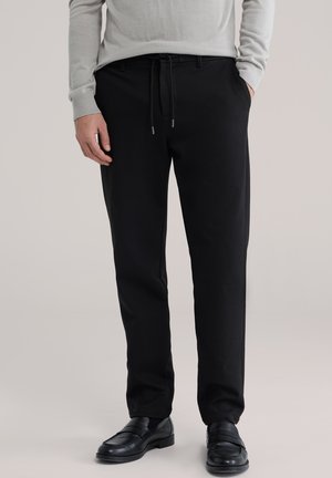 Zwarte, op maat gemaakte broek met een taillekoord, voorzien van een gladde textuur en zijzakken, gecombineerd met zwarte loafers.