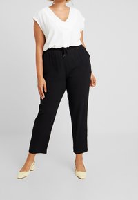 Modelka stojí v bílé blůze s výstřihem do V a černých kalhotách, má na sobě krémové lodičky. Pozadí je neutrální šedé, které zvýrazňuje detaily oblečení.