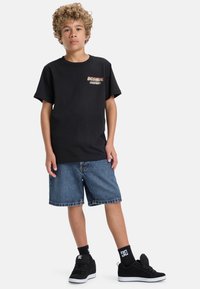 Svart bomullströja med korta ärmar och en flerfärgad "DC SHOE COMPANY" logotyp på bröstet, i kombination med denimshorts och svarta sneakers.
