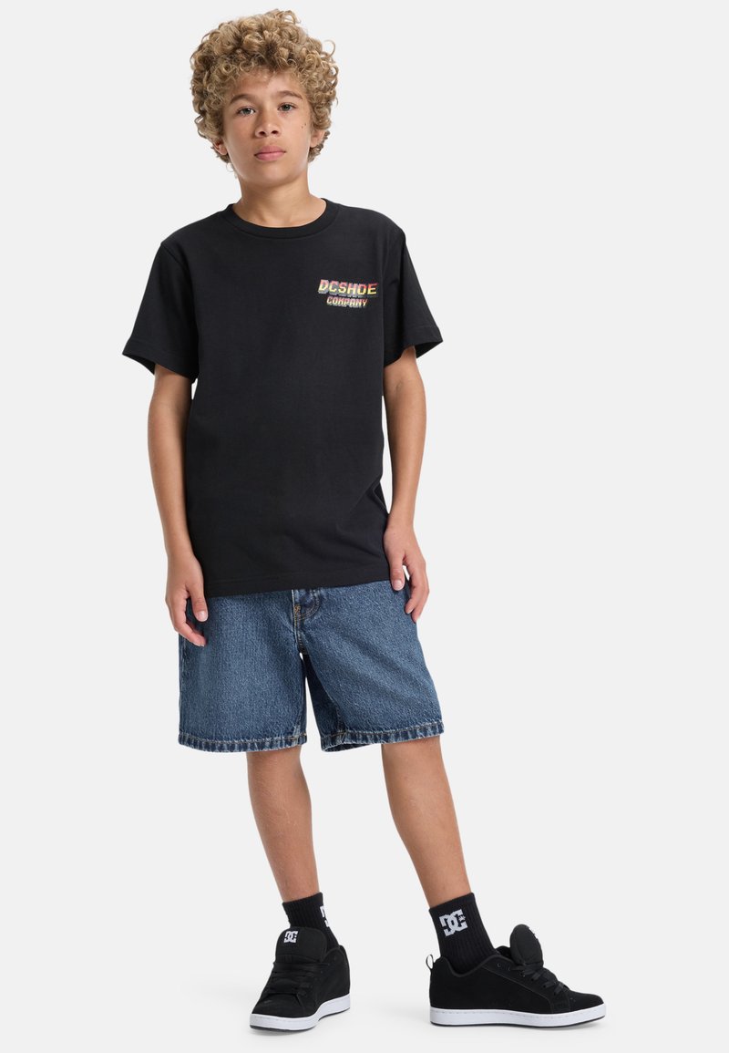 Svart bomullströja med korta ärmar och en flerfärgad "DC SHOE COMPANY" logotyp på bröstet, i kombination med denimshorts och svarta sneakers.
