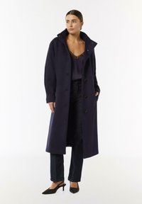 Długi granatowy płaszcz zapinany na guziki, z wysokim kołnierzem i bocznymi kieszeniami, zestawiony z ciemną bluzką i dżinsami oraz czarnymi szpilkami.