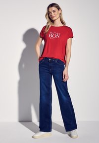 Camiseta corta de manga roja con un gráfico de "BON", combinada con jeans de pierna ancha de color azul oscuro y zapatillas blancas. Textura de algodón suave, corte casual.