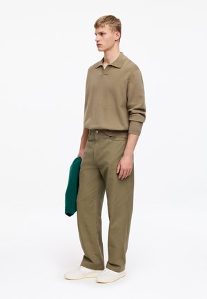 Joven con pantalones verde oliva y camisa beige de manga larga sostiene un suéter verde doblado, de pie contra un fondo blanco liso.
