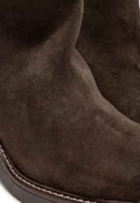 Botas de ante marrón con una textura suave, forma curva y costuras brillantes en contraste a lo largo de la suela. Sin herrajes o patrones notables.