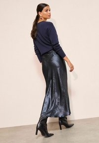 Pulover naval cu mâneci lungi, asortat cu o fustă strălucitoare din satin negru, cizme negre cu vârf ascuțit până la genunchi, privit din spate. Texturi netede și lucioase.