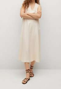 Beige, mouwloze midi-jurk met een V-halsontwerp, vloeiende stof en een ontspannen pasvorm. Gecombineerd met bruine, gespsandalen.