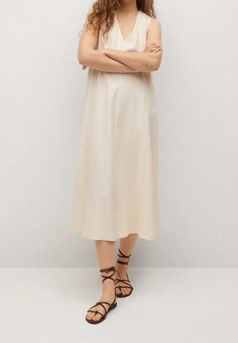 Beige, mouwloze midi-jurk met een V-halsontwerp, vloeiende stof en een ontspannen pasvorm. Gecombineerd met bruine, gespsandalen.