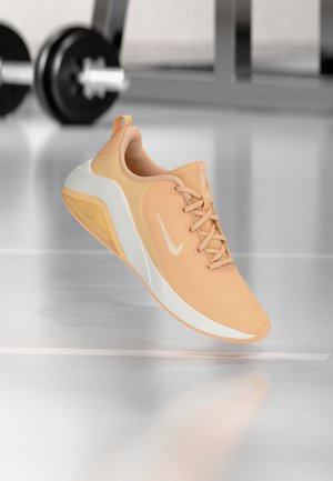 Pantof de alergare Nike de culoare portocaliu deschis, cu logo alb și talpă albă, plutind deasupra unei podele reflectorizante din sală, cu greutăți neclare în fundal.