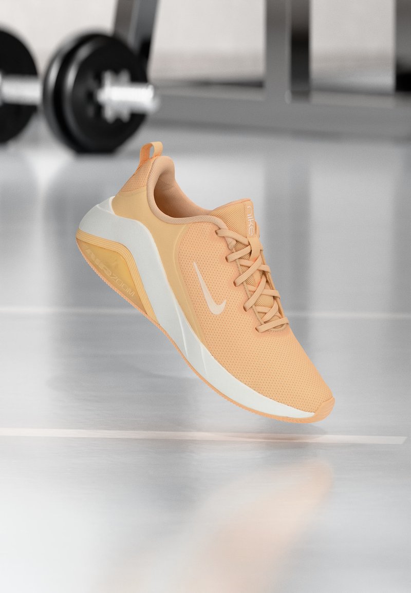 Ανοιχτό πορτοκαλί παπούτσι για τρέξιμο Nike με λευκή λωρίδα και σόλα, να αιωρείται πάνω από ανακλαστικό δάπεδο γυμναστηρίου με θολά βάρη στο φόντο.