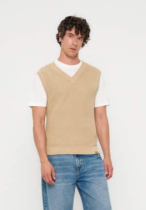 Homme aux cheveux bouclés portant un pull sans manches en tricot beige par-dessus un t-shirt blanc et un jean bleu, debout devant un fond clair uni.