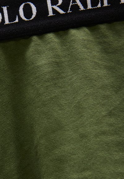 Κοντινό πλάνο πράσινου υφάσματος με μαύρη ζώνη μέσης που φέρει μερικώς ορατό λευκό κεντημένο κείμενο.