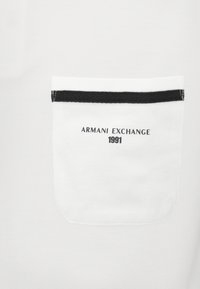 Hvit polo-skjorte med brystlomme med en svart stripe og "ARMANI EXCHANGE 1991" trykt under. Teksturert stoff.