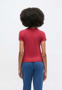 Κόκκινο κοντομάνικο t-shirt με στρογγυλή λαιμόκοψη, λεία υφή, εφαρμοστή γραμμή και εμφανή ελάχιστη ραφή, συνδυασμένο με μπλε φόρμα.