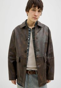 Brown leather jacket with a buttoned front, collar, and two front pockets. Textured surface with a slight sheen, worn over a cream ribbed shirt.