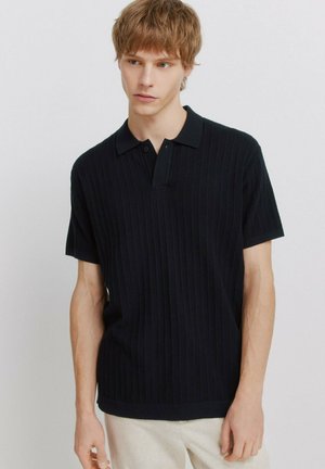 Młody mężczyzna o jasnobrązowych włosach, ubrany w czarną prążkowaną koszulkę polo z krótkim rękawem i jasne spodnie, stojący na tle jednolitego tła.