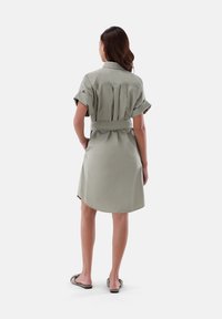 Abito camicia kaki con maniche corte arrotolate, vita con cintura e orlo leggermente curvato. Il tessuto appare liscio con una vestibilità comoda.