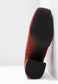 Une chaussure en suédine couleur rouille reposant sur son talon, montrant une semelle noire texturée et un talon bloc, positionnée contre une surface blanche et un mur.