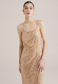 Nude obleka s srebrnimi bleščicami in tankimi naramnicami, prilega se telesu, ima stranski razporek in gladko teksturo, ki odseva svetlobo od številnih bleščic.