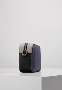 Čtvercová taška vyrobená z námořnického modrého a černého syntetického materiálu, se šedým vrškem, stříbrným zipem a dekoračním kroužkem na boku.