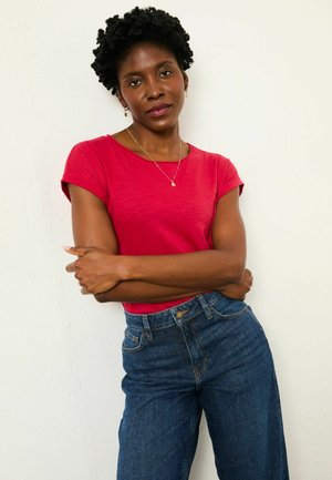 Rotes T-Shirt mit kurzen Ärmeln, kombiniert mit hoch taillierter, dunkelblauer Jeans aus Denim, mit glatter Textur und lockerem Schnitt. Model posiert mit verschränkten Armen.