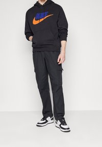 Svart huvtröja med blå och orange Nike-logga, tillsammans med svarta cargo-byxor. Skorna har en svart och vit design. Avslappnad atletisk stil.