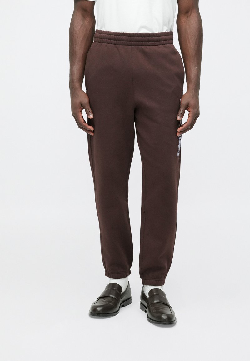 Barna melegítőnadrág elasztikus derékpánttal, oldalzsebekkel és laza szabással. Simogató tapintású, finom márkajelzéssel a lábszárnál.